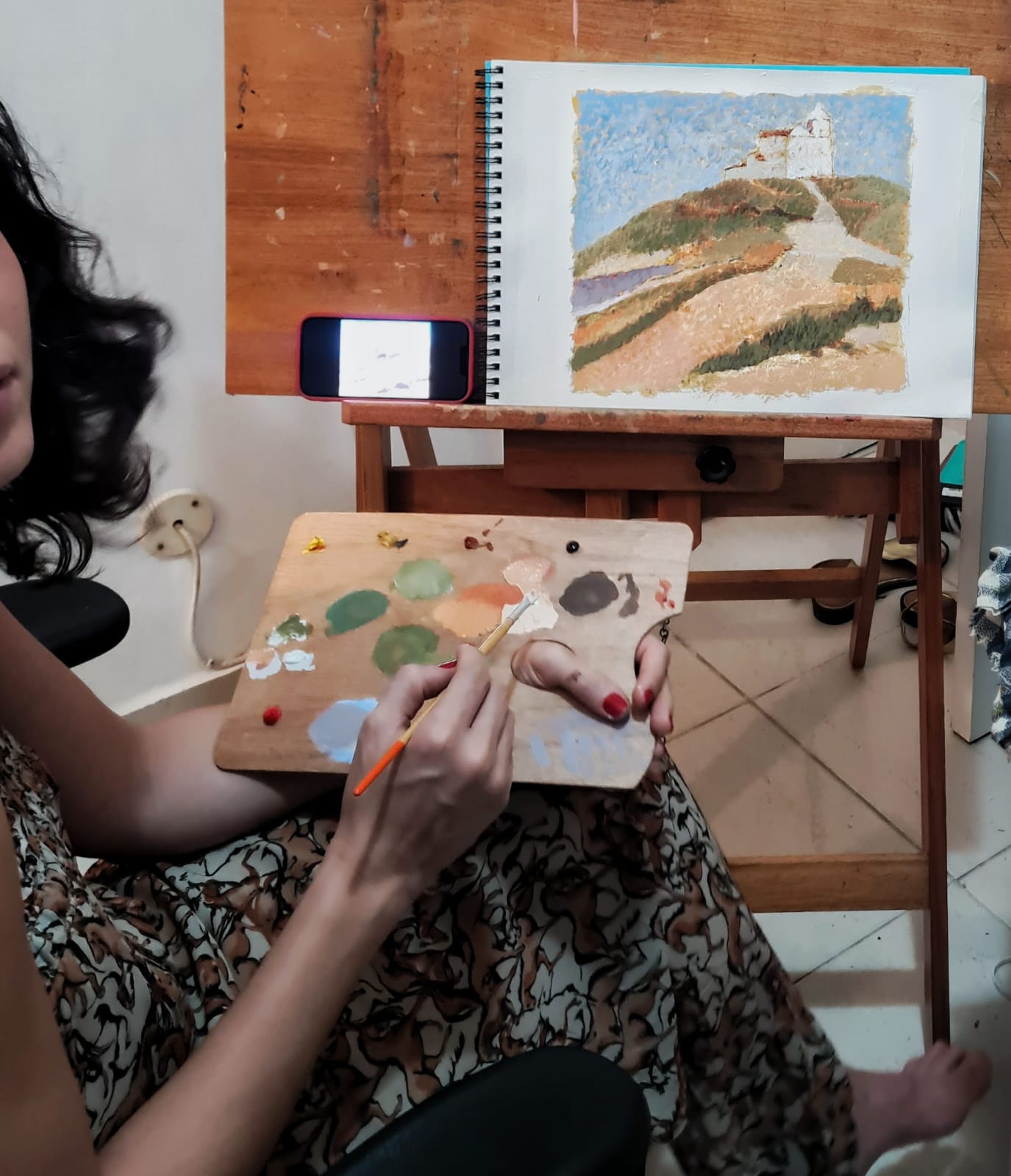 Aluna pintando a óleo uma paisagem com igreja sobre uma colina no atelier de Gabriel Paz em Botafogo, com pincel e paleta em mãos.